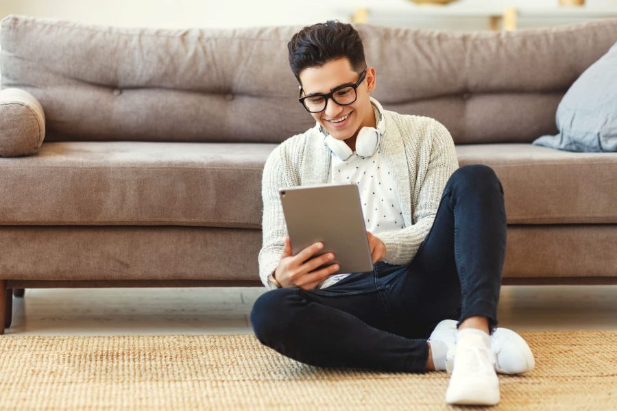Leser:innenführung: Ein junger, gut aussehender Mann sitzt vor einem Sofa und liest auf seinem Tablet.